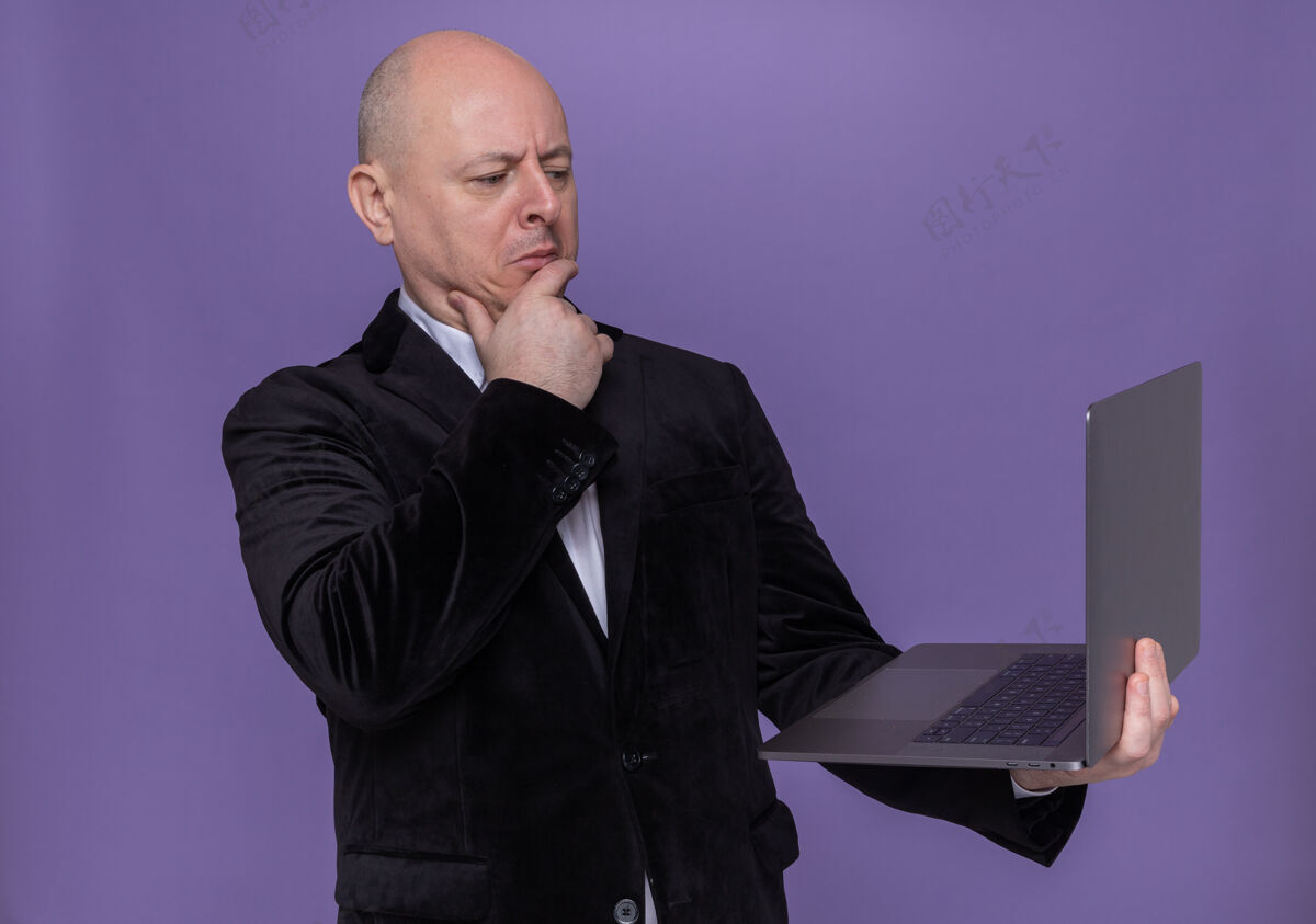 秃头中年光头西装革履拿着笔记本电脑看着屏幕 脸上带着沉思的表情站在紫色的墙上思考思考男人西装