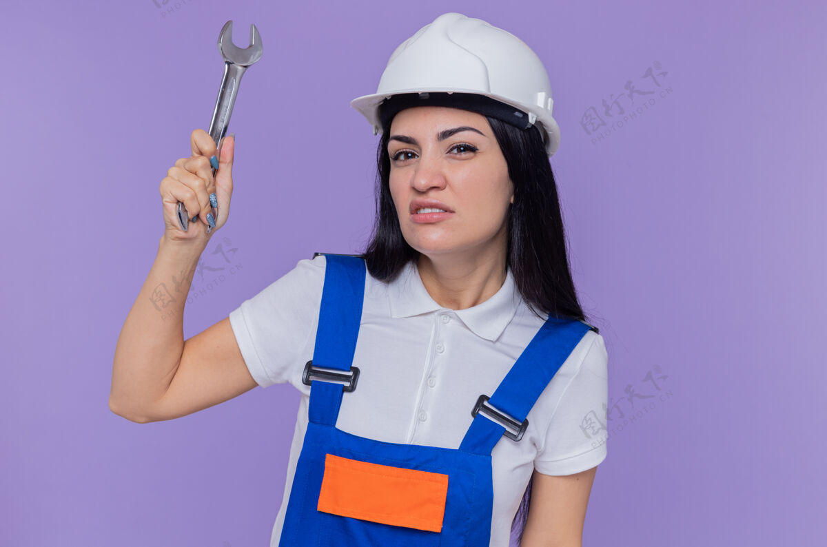 安全身穿施工制服 头戴安全帽 手持扳手的年轻建筑工人站在紫色的墙上 带着怀疑的表情看着前方制服建设者头盔