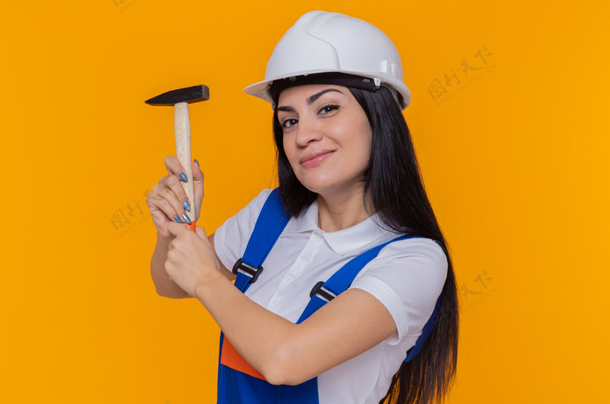 女人身穿施工制服 头戴安全帽 手持铁锤 面带微笑地站在橙色墙壁上的年轻建筑工人安全微笑建筑