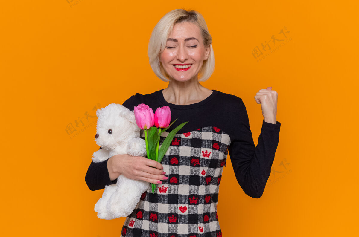 泰迪一位穿着漂亮衣服 手持郁金香和泰迪熊的花束的快乐而兴奋的年轻女子站在橙色的墙上握紧拳头庆祝国际妇女节女人美丽三月