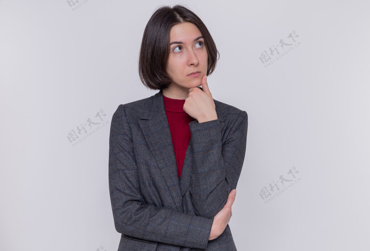 脸一个留着短发 穿着灰色夹克 手放在下巴上 脸上带着沉思的表情 站在白墙上思考的年轻女子短裤站立头发