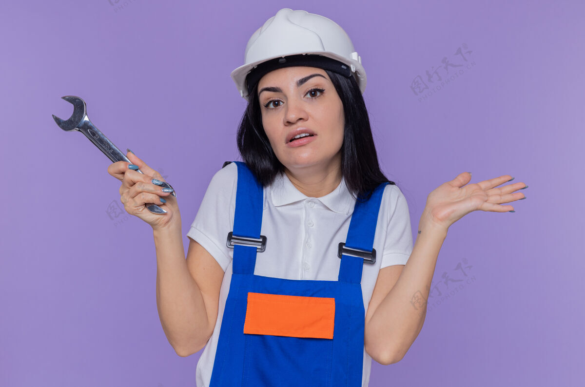 困惑身穿施工制服 头戴安全帽的年轻建筑工人举着扳手 困惑地看着前面 举起手臂站在紫色的墙上 没有回答年轻制服抱着