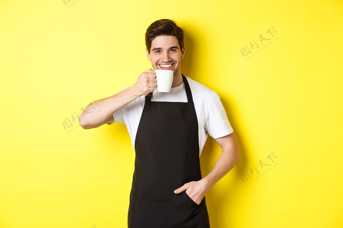 成年人咖啡厅的咖啡师一边喝着咖啡 一边微笑着 穿着黑色围裙 站在黄色的背景上自信烹饪厨师
