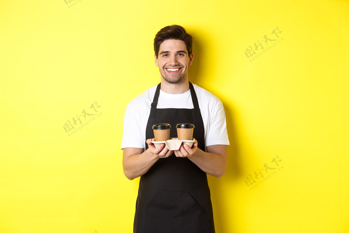 模特帅气的男咖啡师端着外卖咖啡 微笑着 带着秩序 穿着黑色围裙站在黄色背景下咖啡表情年轻