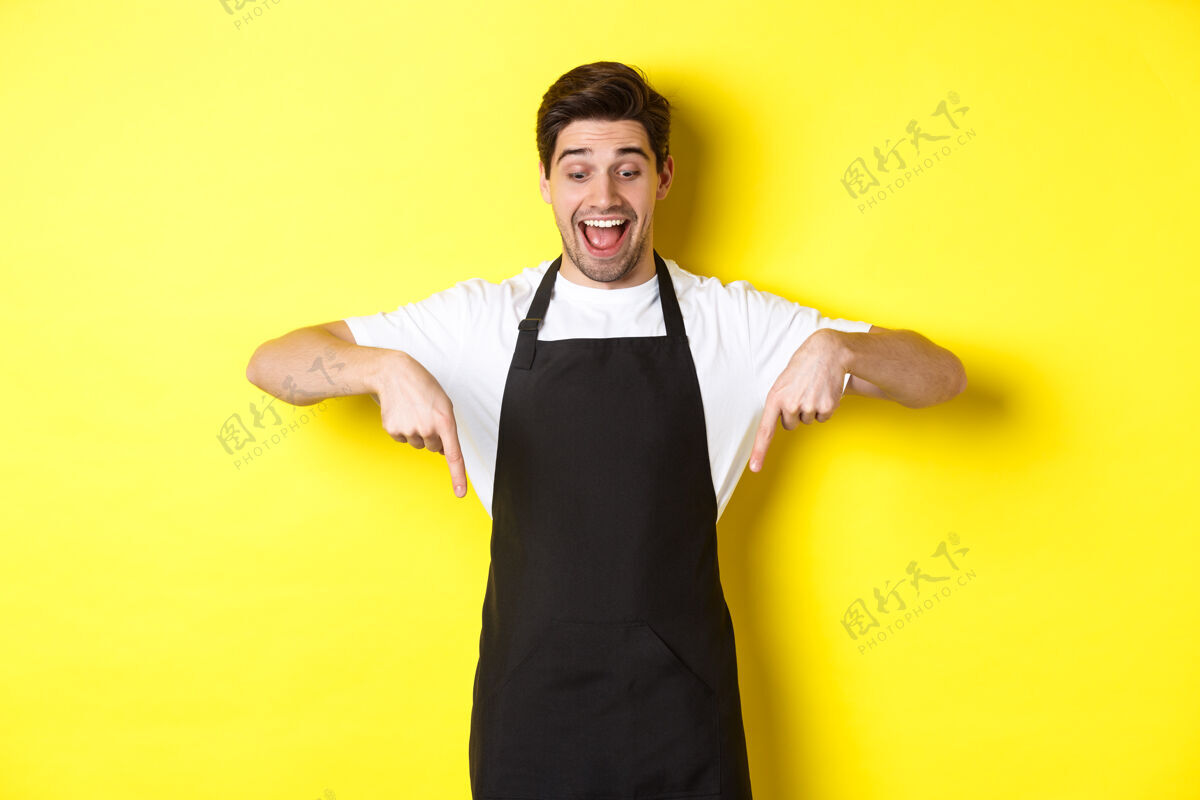年轻穿着黑色围裙的兴奋的侍者手指向下 查看促销优惠 站在黄色背景上帅气烹饪模特