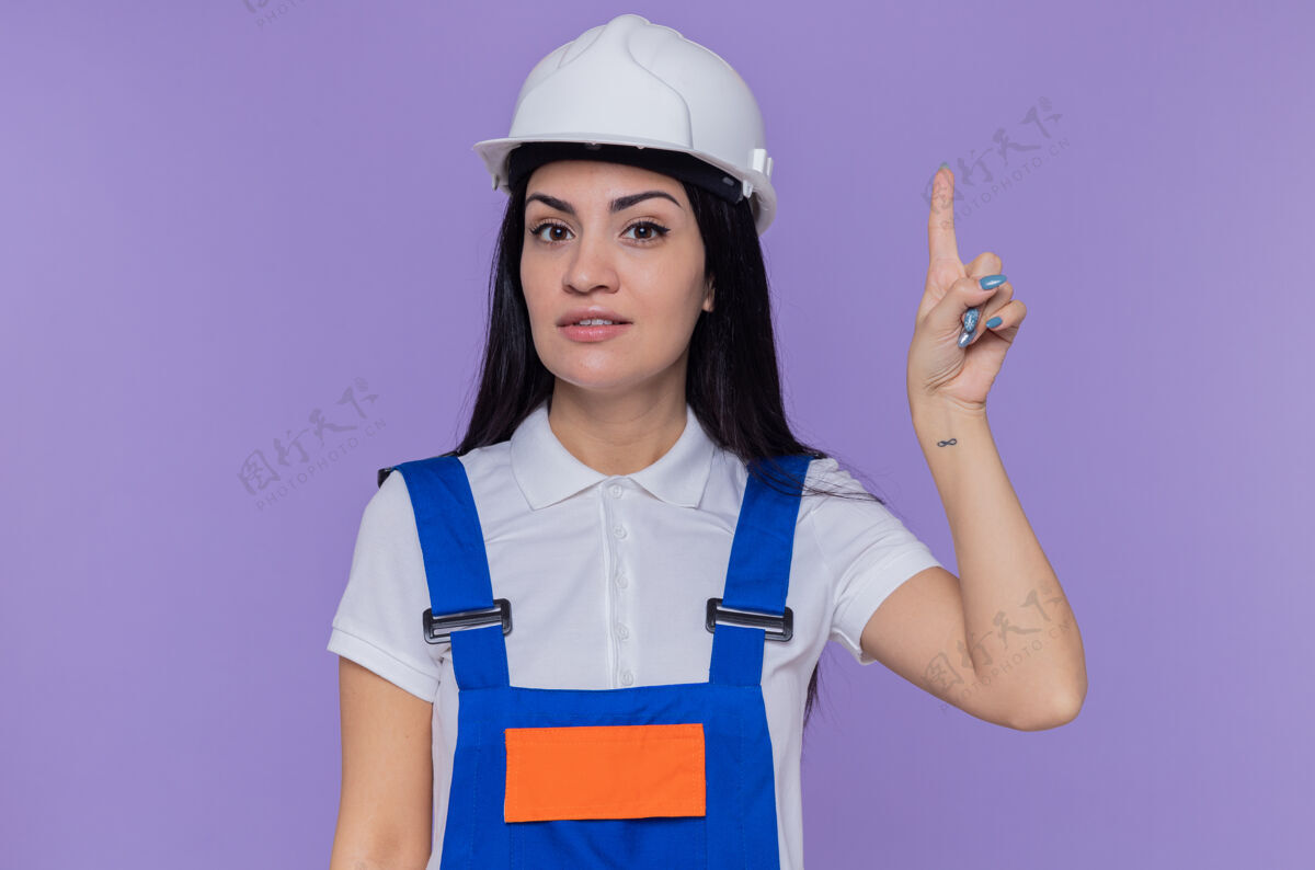 显示身穿施工制服 头戴安全帽的年轻建筑工人站在紫色的墙上 面带微笑地看着前方 露出食指 有着伟大的新想法目录手指建筑