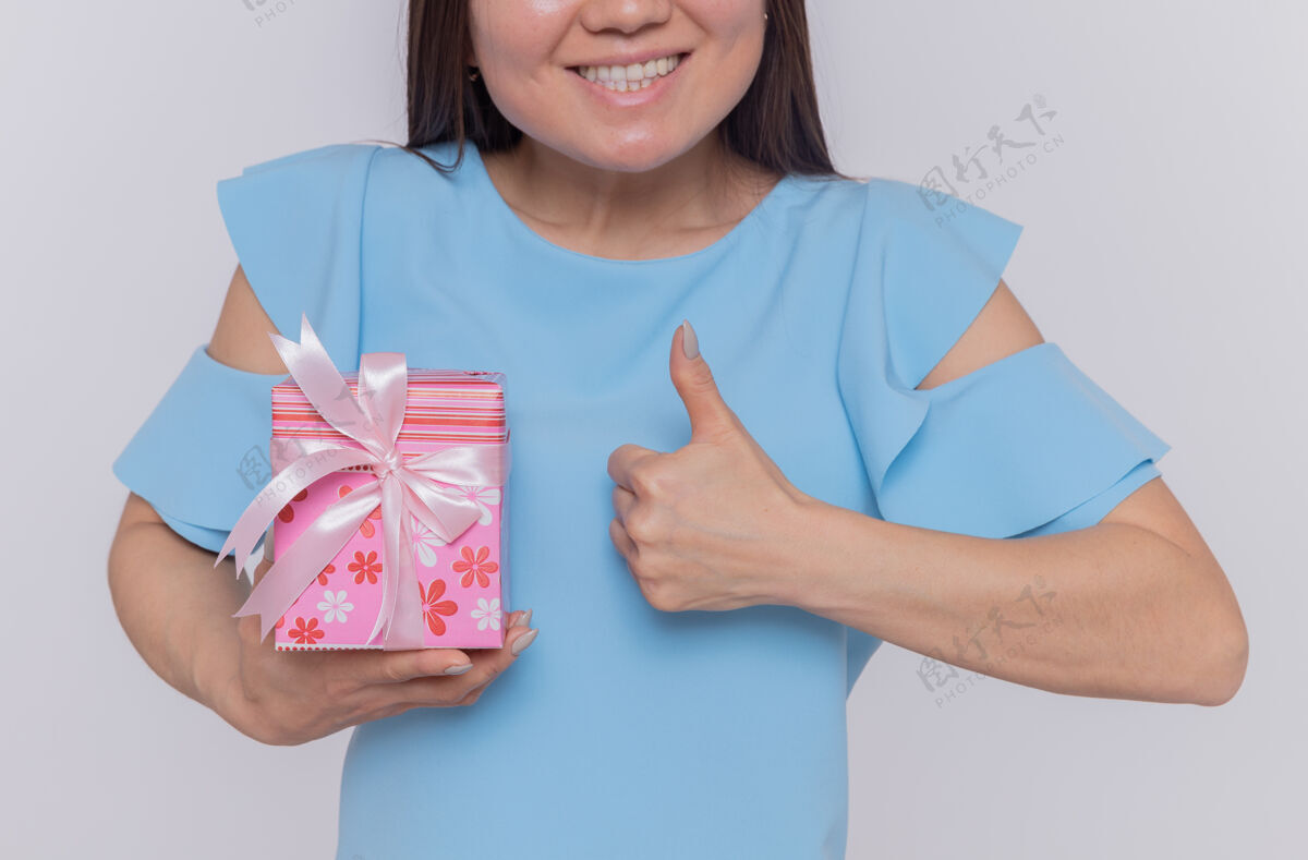 快乐快乐的亚洲女人穿着蓝色的衣服拿着礼物微笑着竖起大拇指站在白色的墙上庆祝国际妇女节庆祝站立服装