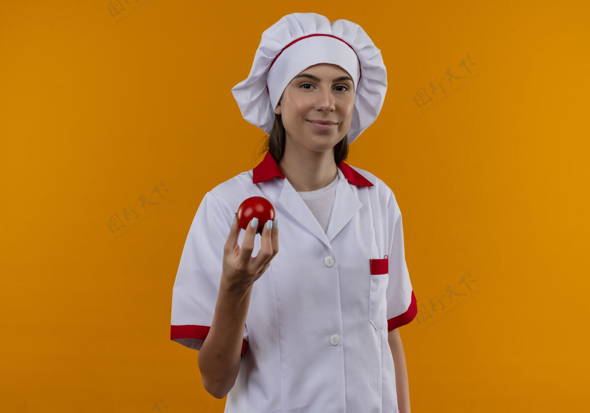 西红柿穿着厨师制服的年轻快乐的白人厨师女孩拿着西红柿看着橙色的相机 留着复印空间副本烹饪制服