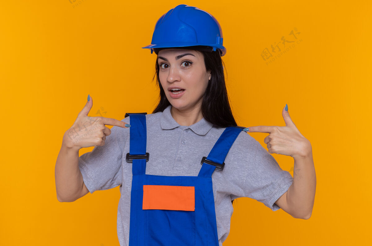 惊喜身穿建筑制服 头戴安全帽的年轻建筑工人面带微笑 看着自己站在橙色的墙上 既高兴又惊讶微笑头盔制服