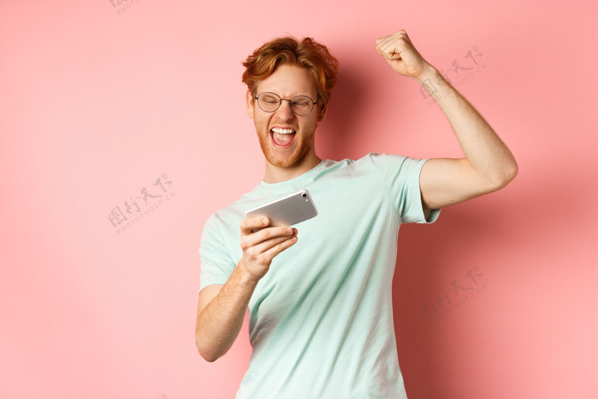 姜快乐的红发人在手机视频游戏中获胜 举起手 高兴地喊“是” 庆祝胜利 看着智能手机 站在粉色背景上男T恤人