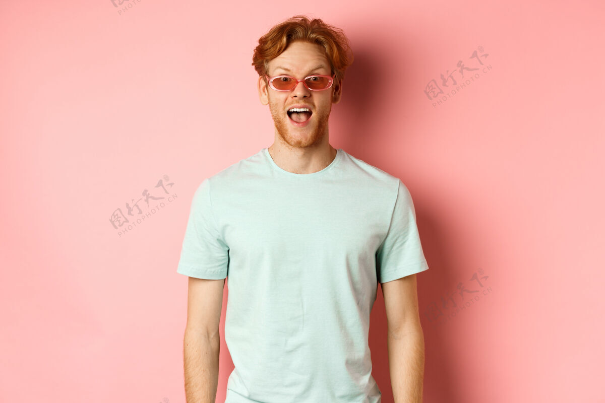 男人度假时惊讶的红发男人 戴着太阳镜 穿着夏季t恤 张开嘴说“哇 惊讶” 站在粉色背景下人脸年轻