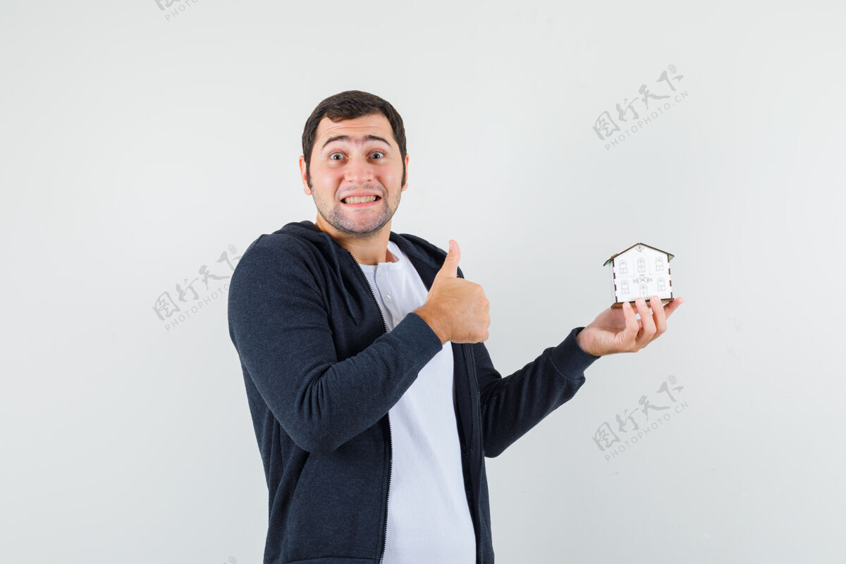 肖像年轻人拿着房子模型和显示拇指在白色t恤和拉链前黑色连帽衫 看起来乐观正面视图房子前面姿势