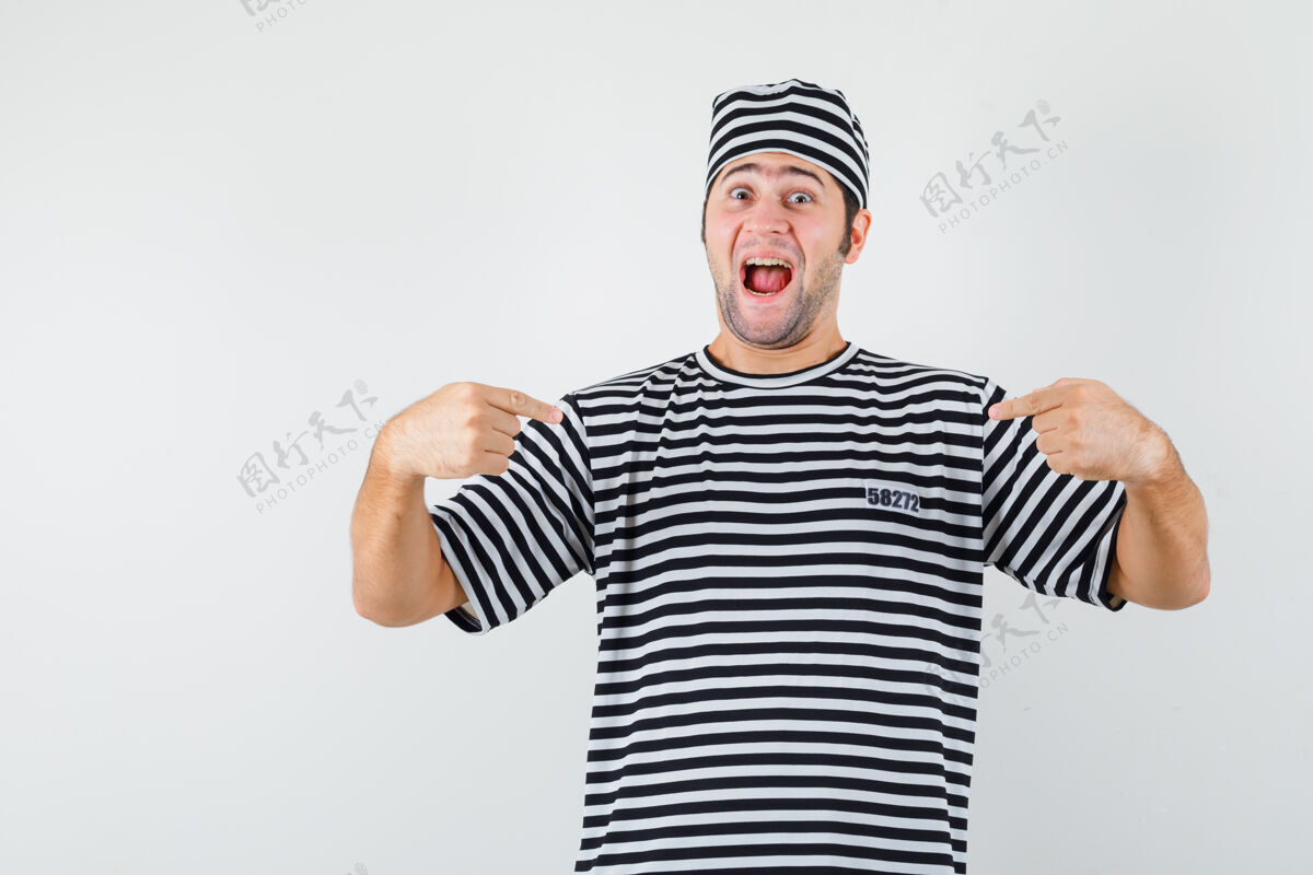 肌肉年轻男子指着自己的t恤衫 帽子 看起来很快乐前视图T恤男人男性
