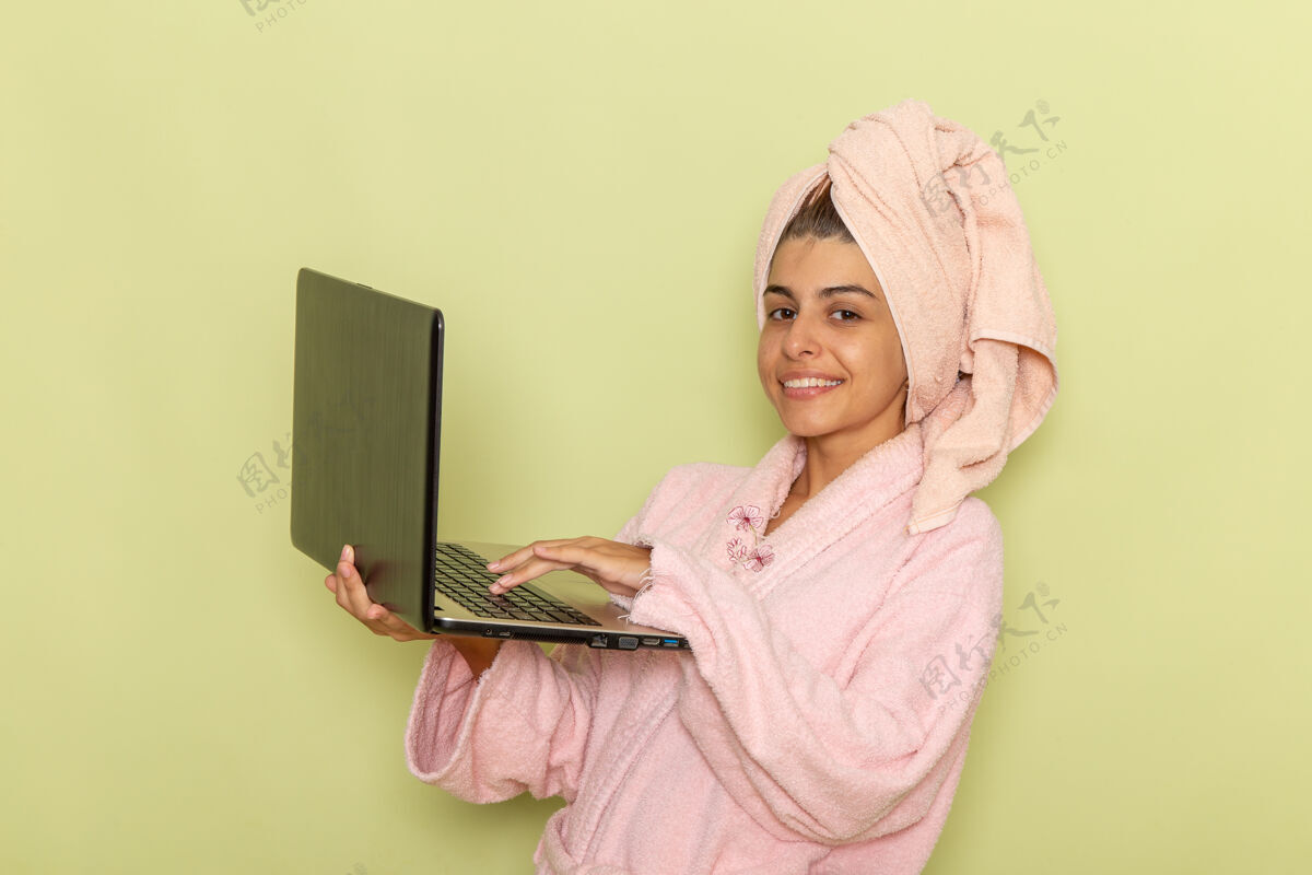 漂亮前视图穿着粉红色浴袍的年轻女性在绿色表面上使用笔记本电脑衣服自我照顾年轻女性