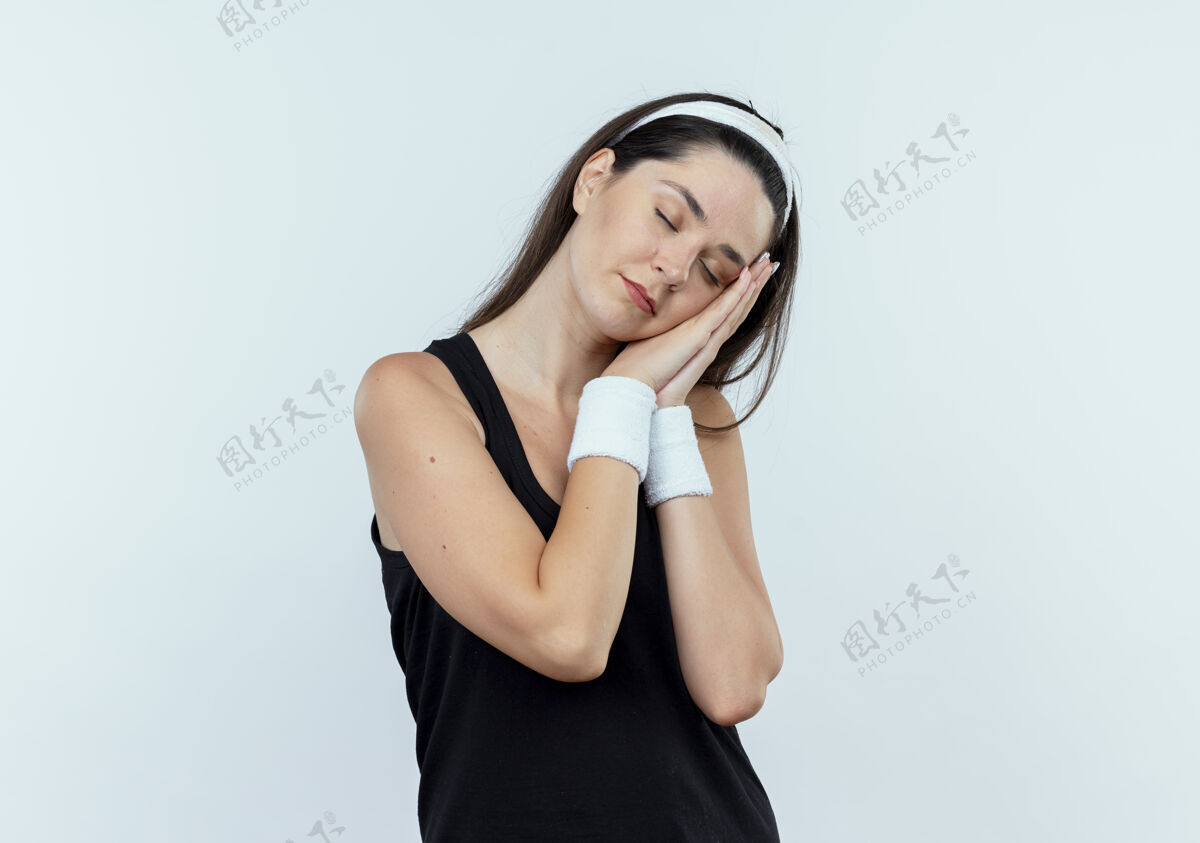 睡眠戴着头巾的年轻健身女士做着睡觉的姿势 手掌靠在手掌上 闭着眼睛站在白色的墙上站立健身手掌