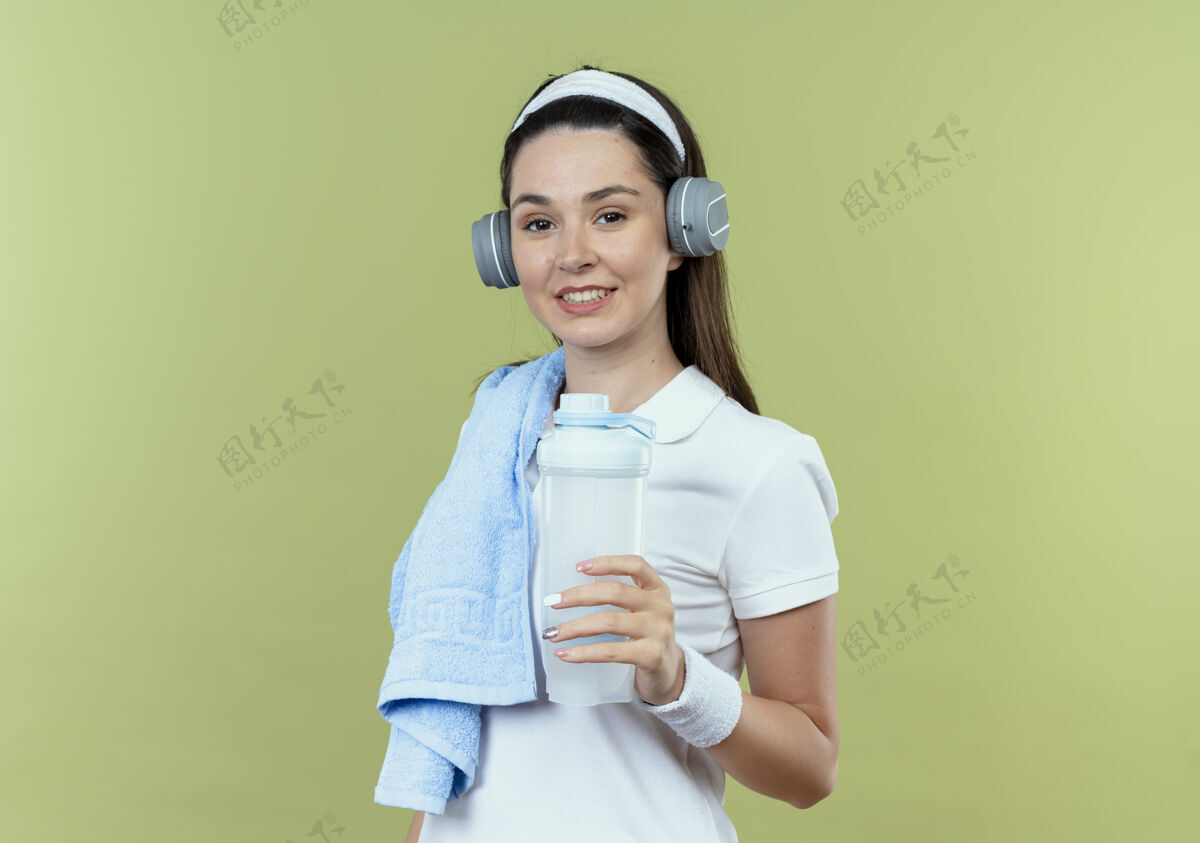 耳机戴着头巾 戴着耳机 肩上扛着毛巾 手里拿着一瓶水 微笑着站在轻墙上的年轻健身女士抱着年轻微笑