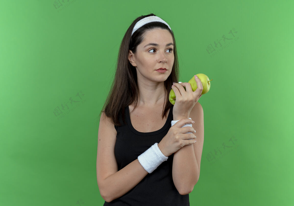 年轻的女人戴着头巾的年轻健身女手拿着青苹果 严肃的脸站在绿色的墙上望向一边把头戴在一边