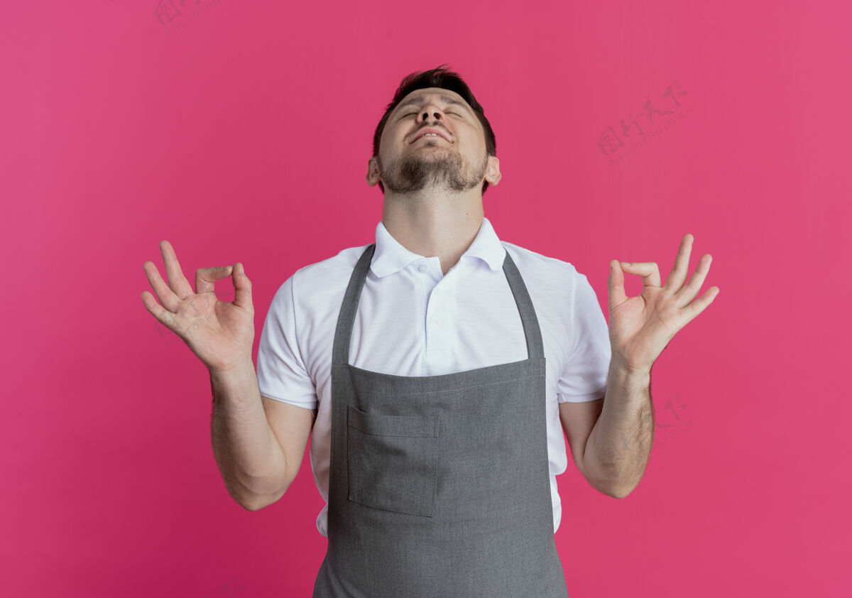 手势围裙上的理发师正在放松 闭着眼睛站在粉红色的墙上做着冥想的手势关闭男人冥想