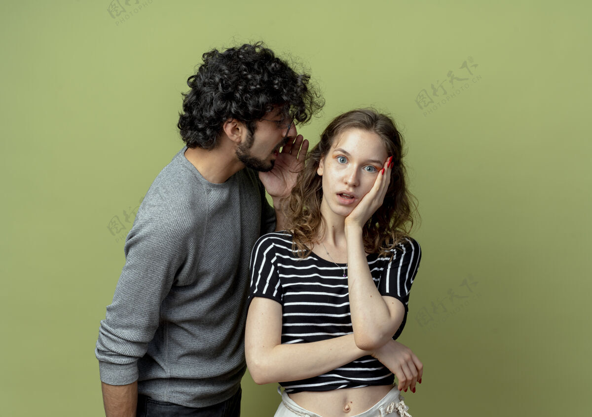 女人年轻漂亮的情侣男女 男人在浅绿色的墙上对女友小声说秘密或有趣的闲话流言蜚语男人光
