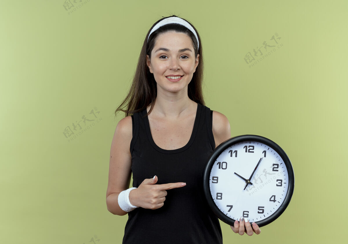 女人戴着头巾的年轻健身女士拿着挂钟 手指指着它微笑着站在明亮的墙上墙光抱着