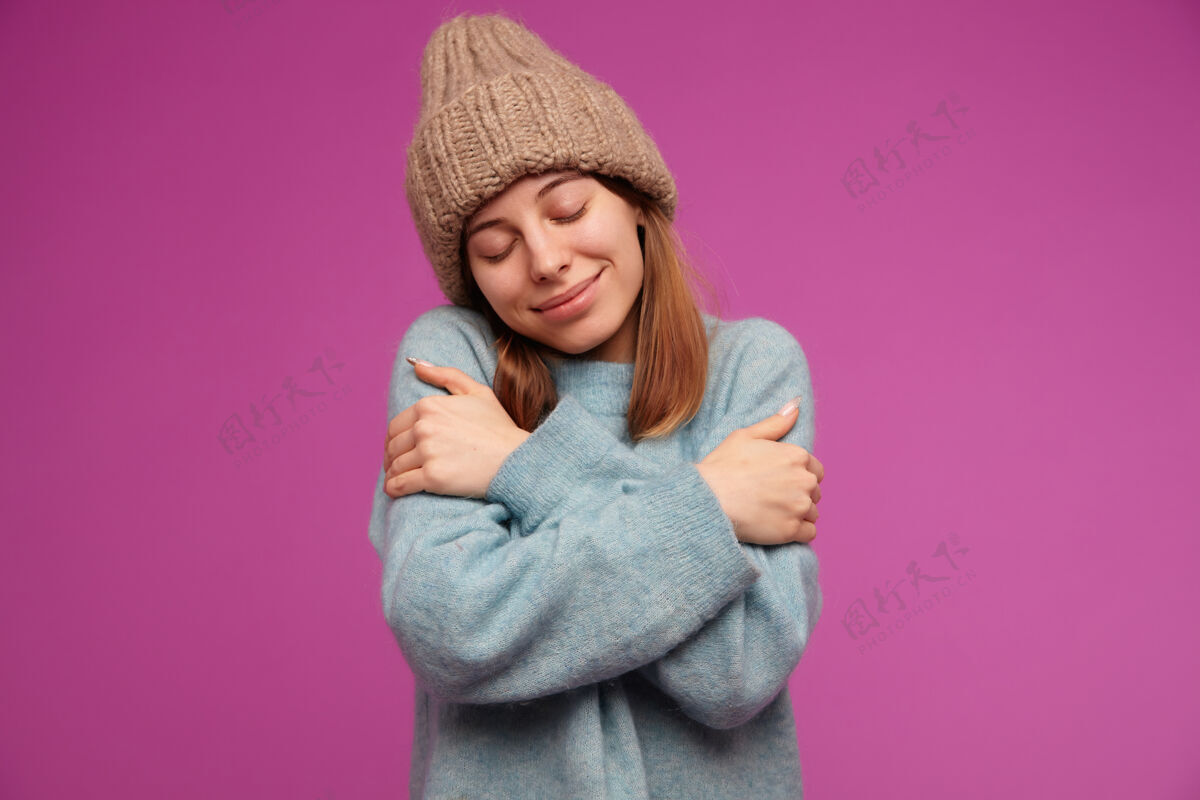 完美十几岁的女孩 快乐的女人 深色长发 穿着蓝色毛衣 戴着针织帽子 拥抱自己 在紫色的墙上感觉温暖和舒适黑发皮肤休闲