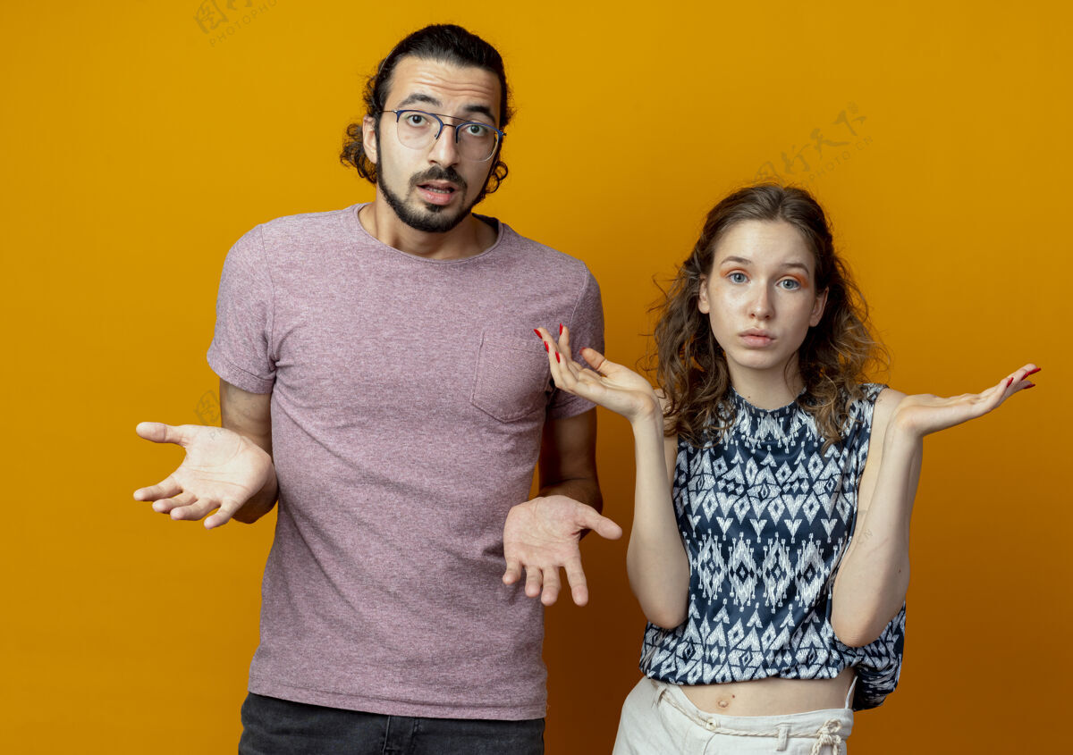 男人年轻漂亮的夫妇男人和女人困惑和不确定没有答案 跳到一边站在橙色墙困惑不确定手臂