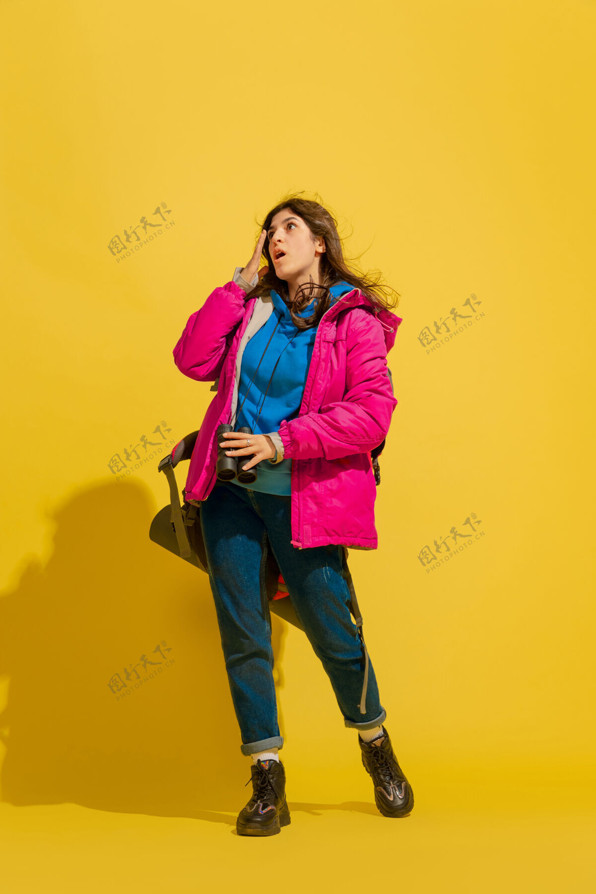 肖像黄色摄影棚墙上 一个带着包和望远镜的快乐的年轻旅游女孩的画像多彩方式指向