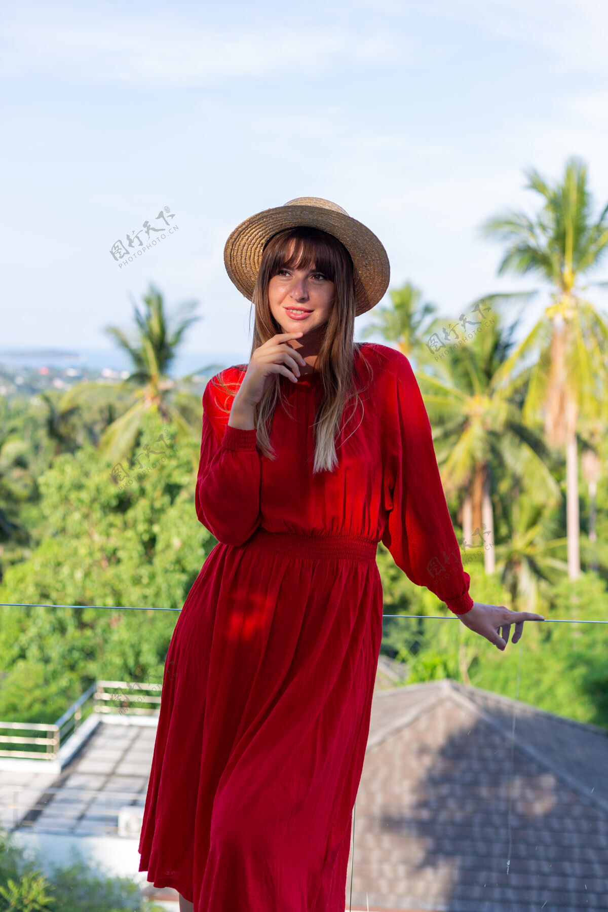 夏季穿着红色夏装 戴着草帽 在阳台上享受热带海景和棕榈树的快乐女人放松年轻站立