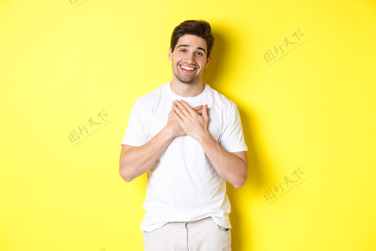 自信一个穿着白色t恤的充满感激的帅哥 手牵着手 开心地微笑着 表达感激 感谢某事 站在黄色的背景上谢谢男人成年人