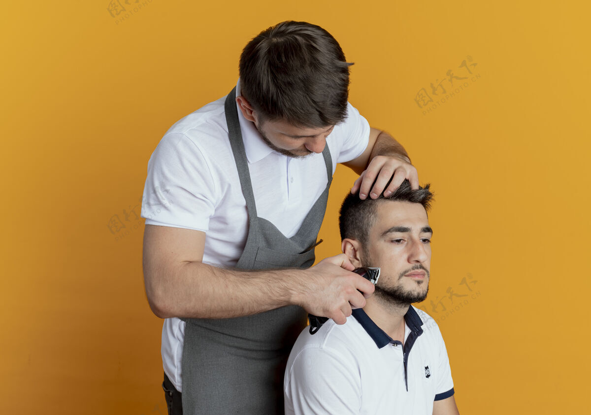 男人穿着围裙的理发师用满意客户的胡须修剪器在橘色墙上刮胡子满意胡须顾客