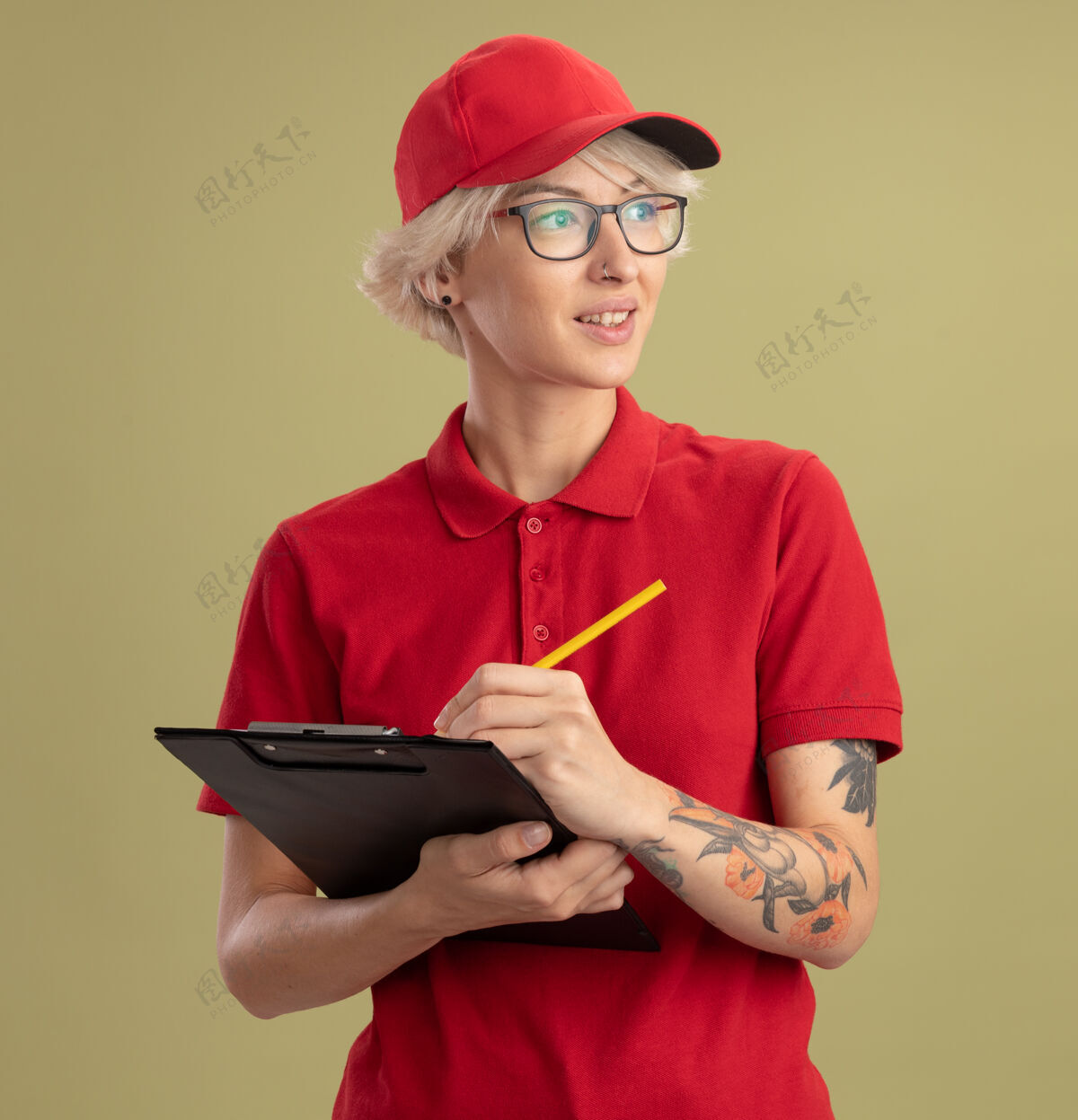 戴着年轻的送货员身穿红色制服 头戴帽子 戴着带剪贴板的眼镜和铅笔 脸上带着微笑站在绿色的墙上脸眼镜女人