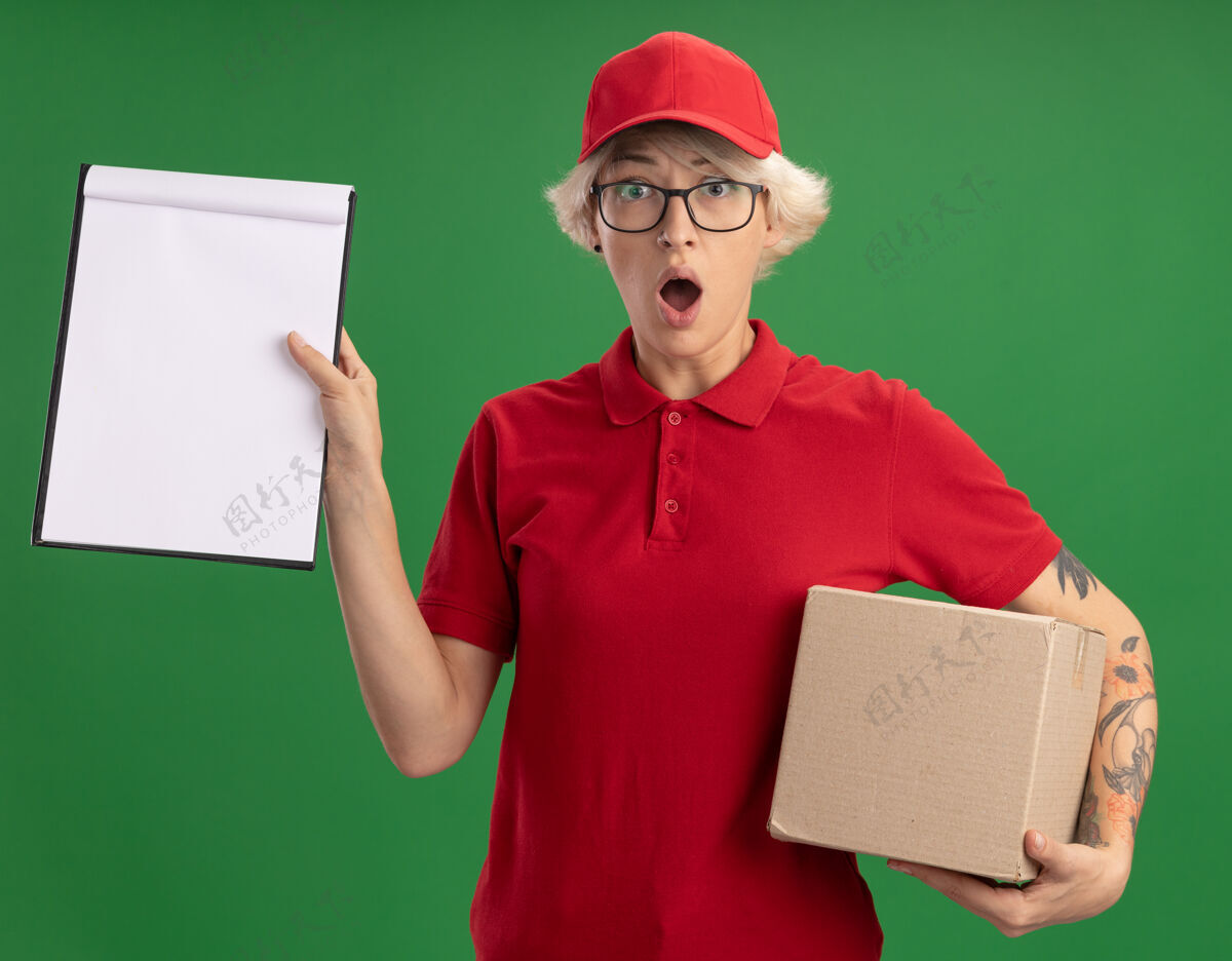 穿身穿红色制服 戴着帽子 戴着眼镜的年轻女送货员站在绿色的墙上 手里拿着一个纸板箱 上面有空白页的剪贴板 忧心忡忡 大吃一惊帽子年轻眼镜