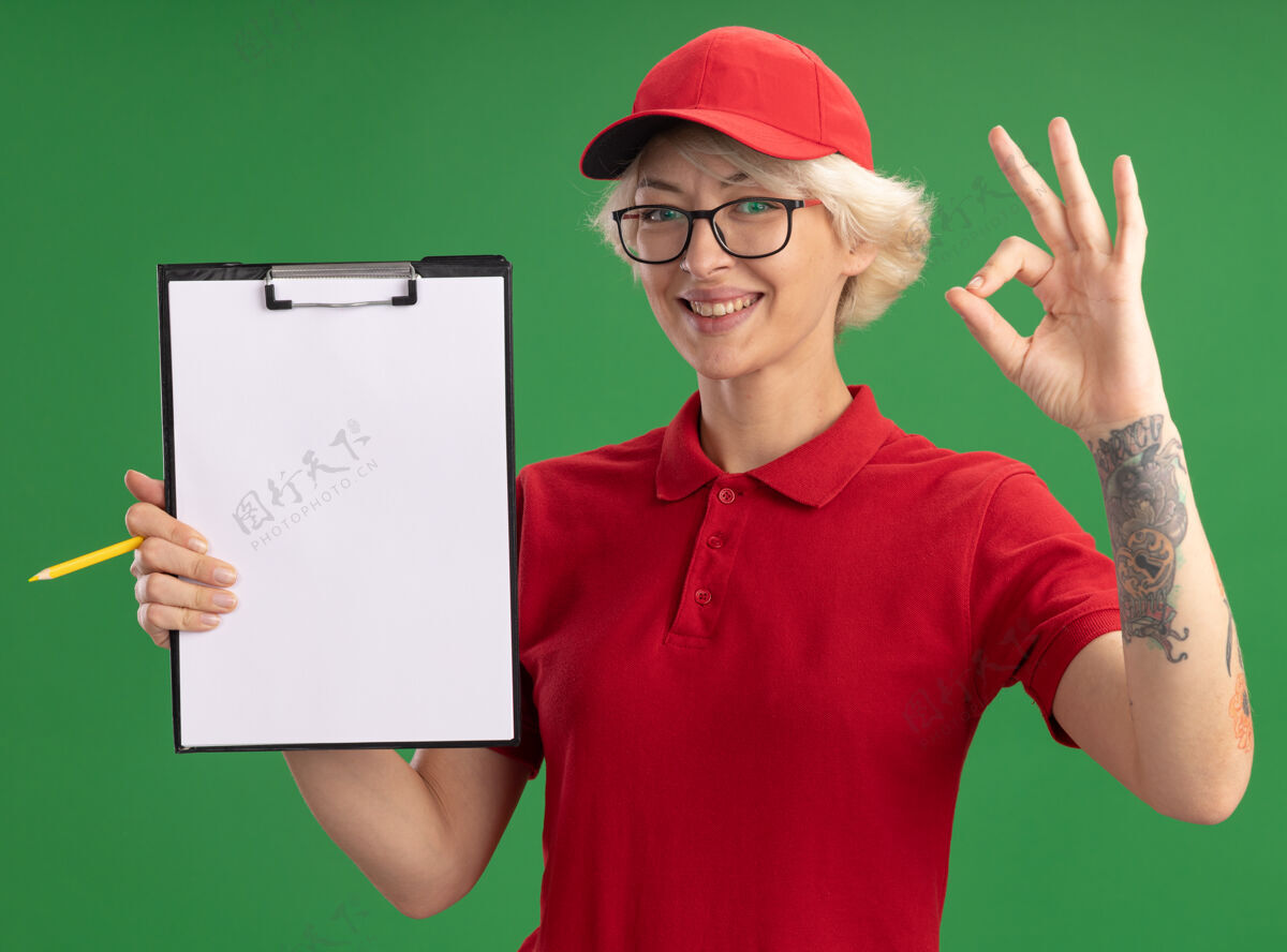 剪贴板身穿红色制服 戴着帽子的年轻送货员戴着一副眼镜 上面有空白页的剪贴板和铅笔 她愉快地微笑着 绿色的墙上挂着“ok”的牌子微笑站穿着