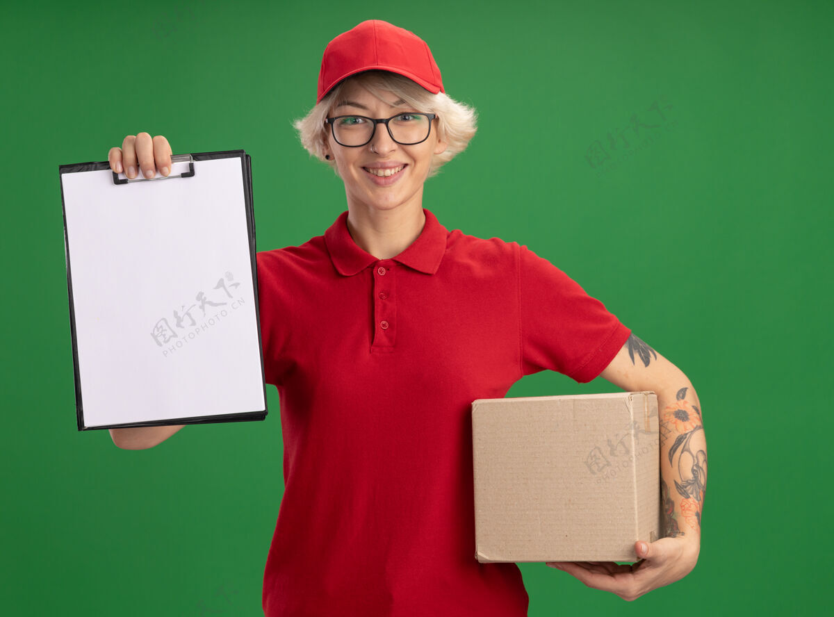 递送身穿红色制服 戴着帽子 戴着眼镜的年轻送货员站在绿色的墙上 手里拿着一个纸板箱 上面有空白页的剪贴板 脸上洋溢着愉快的笑容站立制服帽子