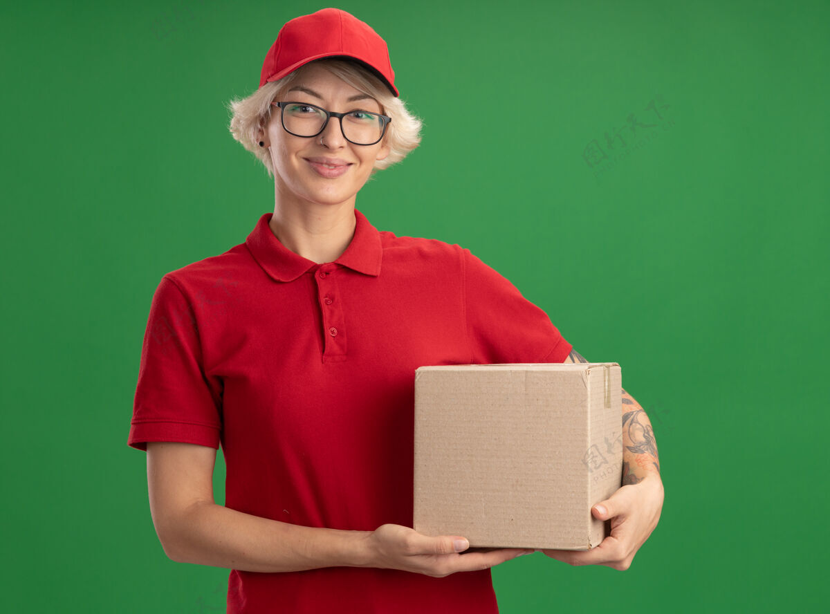 年轻年轻的送货员身穿红色制服 戴着帽子 戴着眼镜 手里拿着纸板箱 站在绿色的墙边 面带微笑 自信满满眼镜信心帽子