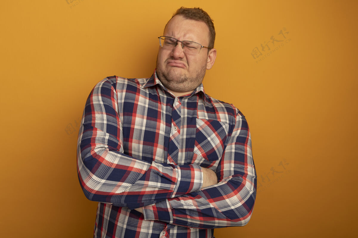 站一个戴眼镜 穿格子衬衫的男人不满意地交叉双臂站在橙色的墙上皱着眉头的脸男人脸交叉