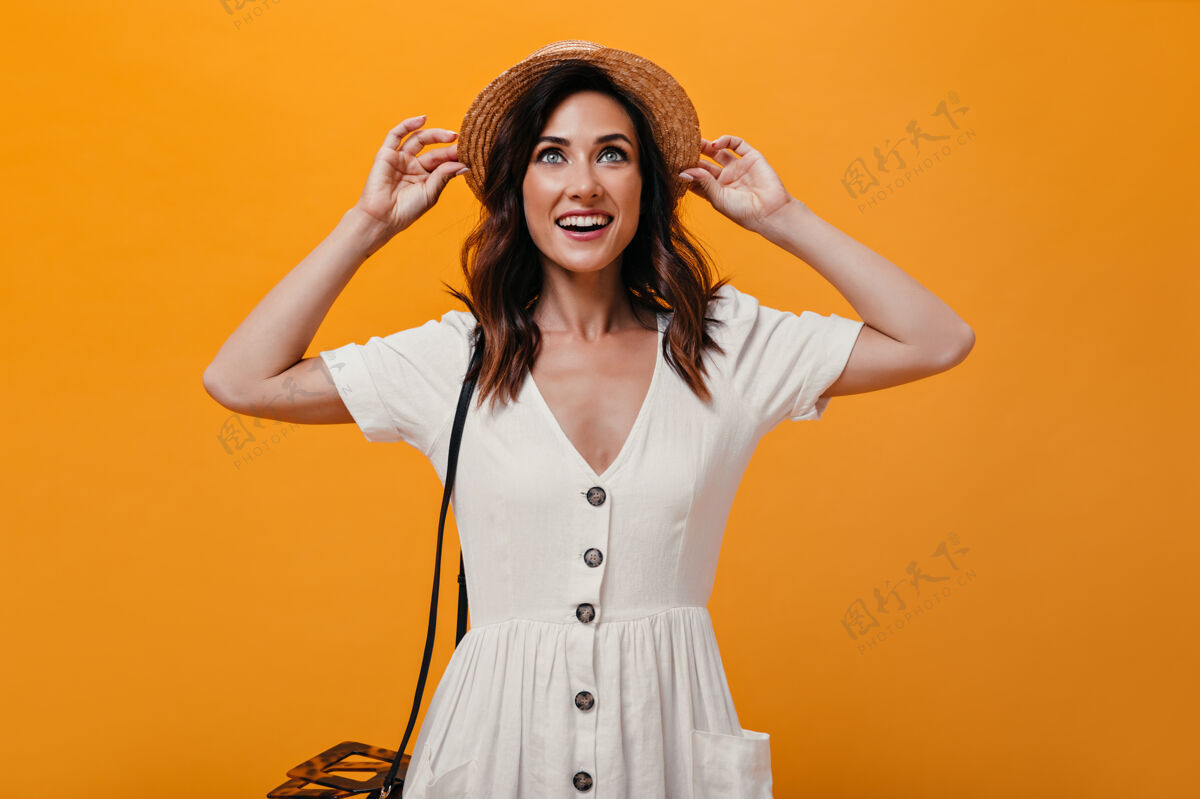 思考美丽的女孩拿着帽子 在橙色的背景上微笑迷人的女人 黑色短发 穿着白色连衣裙 摆姿势微笑着全景沉思休闲