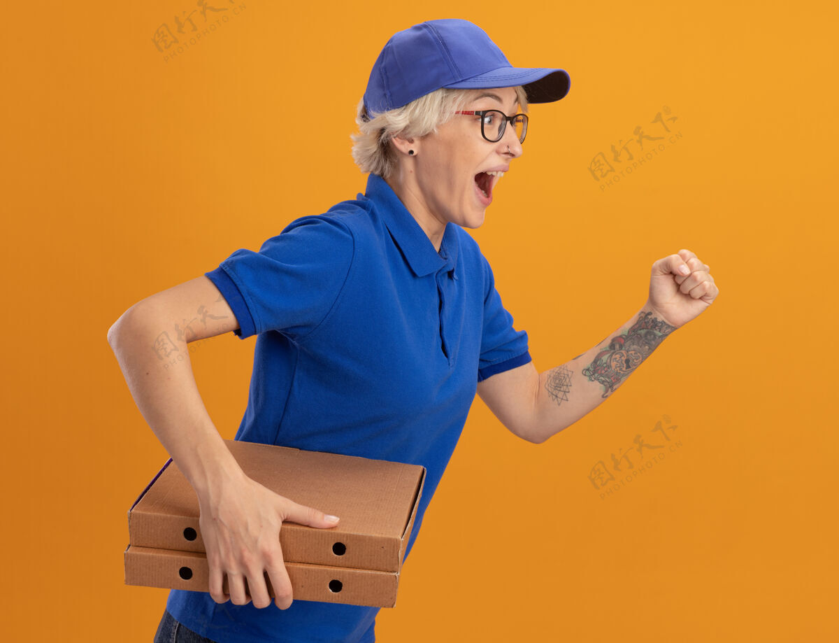 匆忙身穿蓝色制服 头戴鸭舌帽的年轻送货员兴奋地跑过去 越过橘色的墙壁为顾客送比萨饼盒送货女人制服