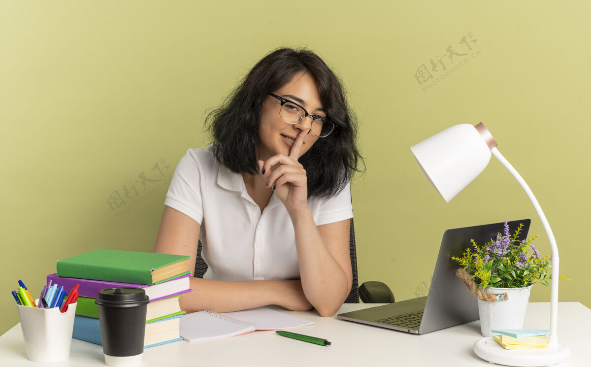 学校年轻自信漂亮的白人女学生戴着眼镜坐在书桌旁拿着学习工具手势安静地把手指放在嘴上隔离在绿色空间和复印空间绿色穿坐