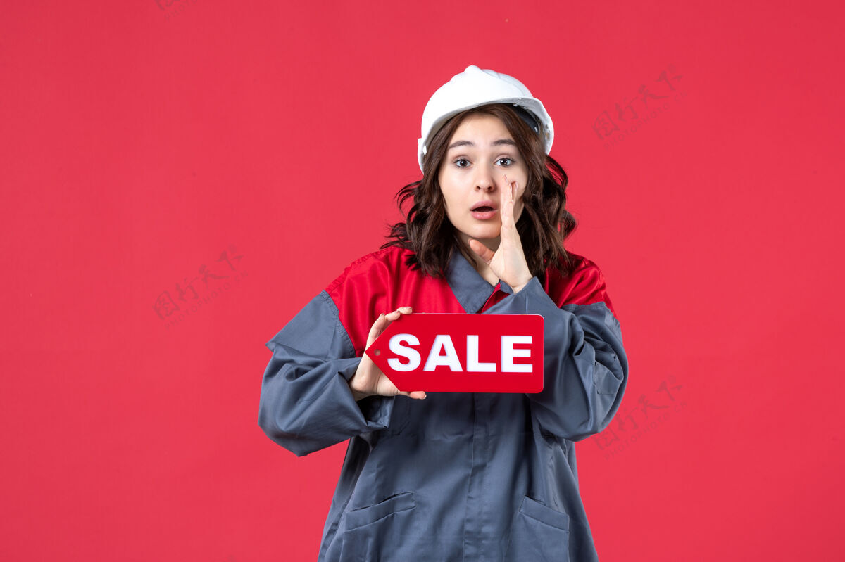 微笑正面是穿着制服戴着安全帽的女工的近景 展示着销售图标 并在隔离的红墙上呼唤着某人肖像前面女工