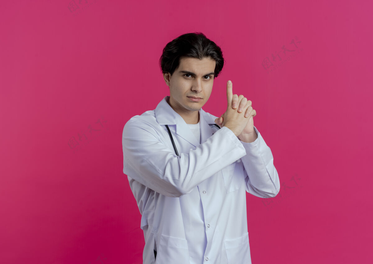 穿自信的年轻男医生穿着医用长袍和听诊器做手枪手势隔离在粉红色的墙上与复制空间手枪年轻听诊器