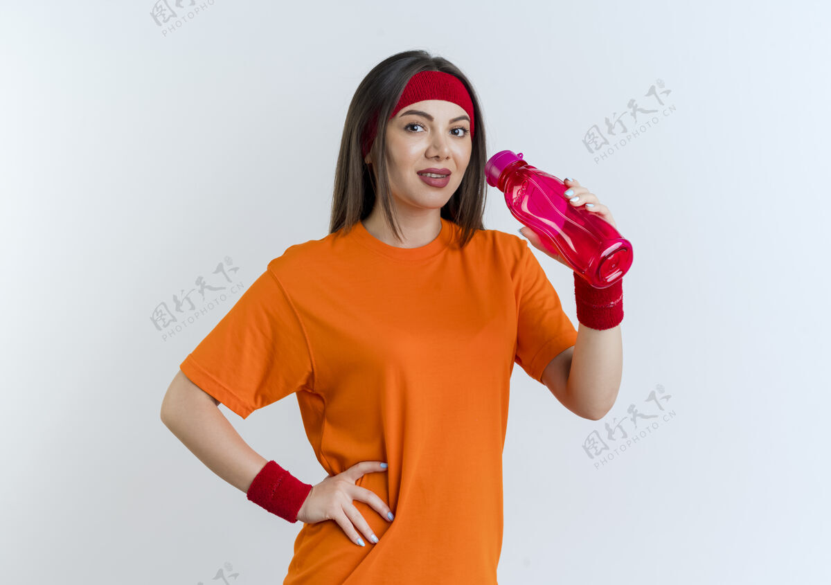 运动喜欢运动的年轻女子戴着头带和腕带拿着水瓶 手放在腰上 隔离在白色墙壁上 留有复制空间手女人年轻