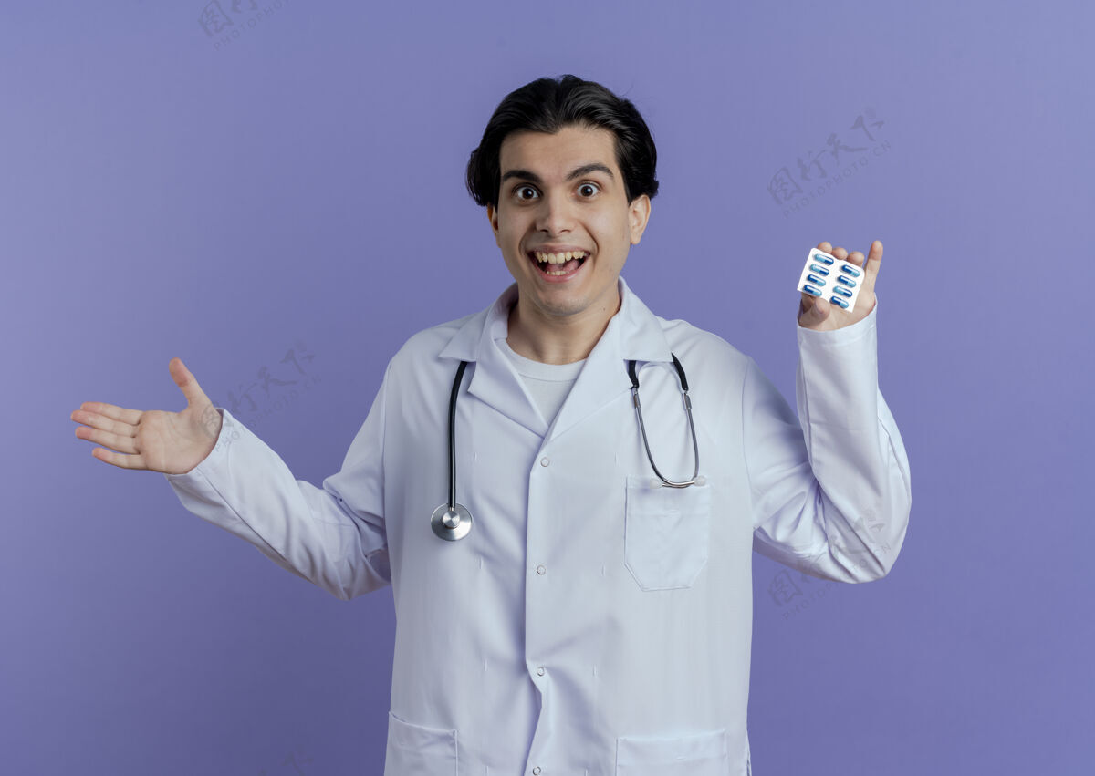 医生令人印象深刻的年轻男医生穿着医用长袍和听诊器显示胶囊包显示空手隔离在紫色的墙壁上穿医学听诊器