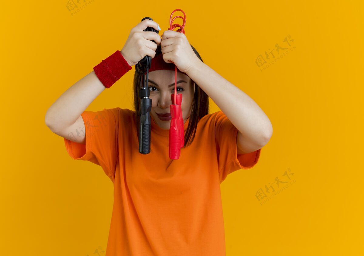 请戴着头带和腕带的年轻运动型女士高兴地拿着跳绳 让他们在橘色墙壁上的脸与复制空间隔离头带橙色抱着