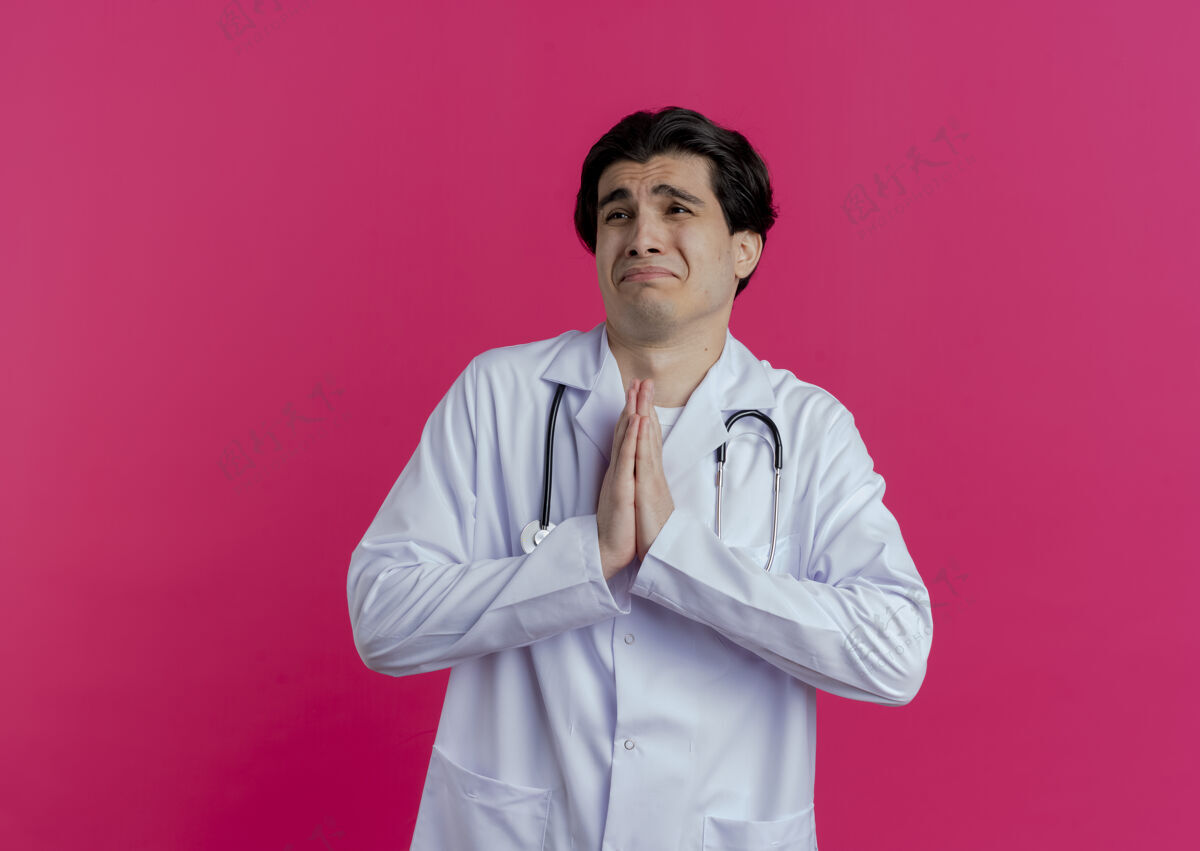 听诊器悲伤的年轻男医生穿着医用长袍和听诊器看着一边双手合十乞求隔离在粉红色的墙上有复印空间医生年轻手