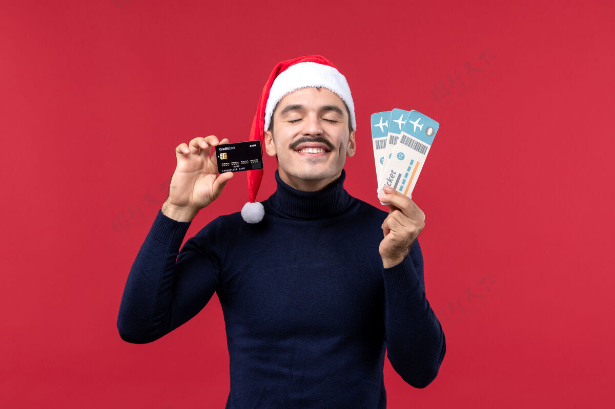 微笑正面图年轻男子手持红色背景的机票和银行卡肖像银行圣诞节