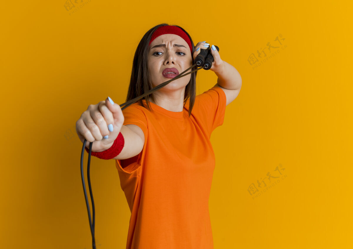 腕带皱着眉头戴着头带和腕带的年轻运动女性用跳绳练习看着它孤立在橙色的墙上 有着复制的空间穿头带运动