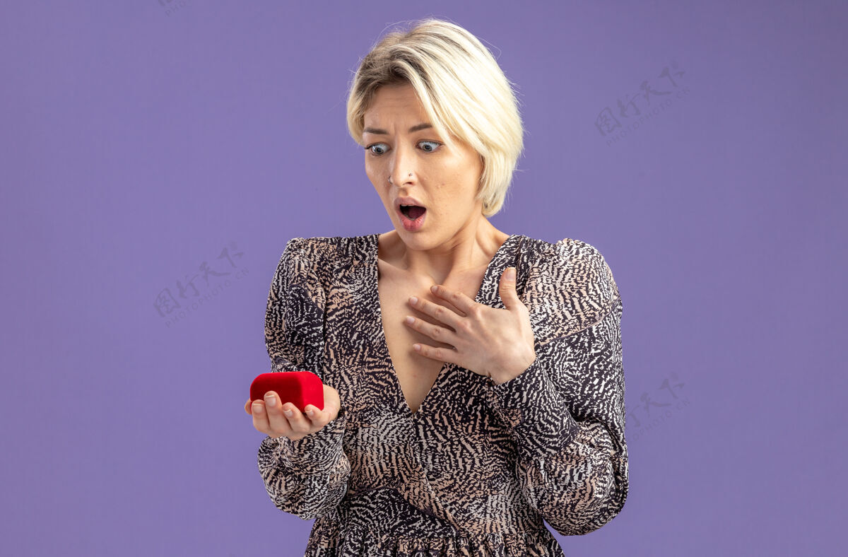举行年轻漂亮的女人穿着礼服拿着红色的订婚戒指看着它惊讶和惊喜的情人节概念站在紫色的墙上年轻情人节期待
