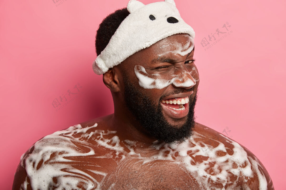 水滑稽的大胡子男人眨着眼睛 站在满是泡沫的地方 在浴室里洗澡 戴着柔软的白色头带 隔离在粉红色的墙上卫生室内新鲜