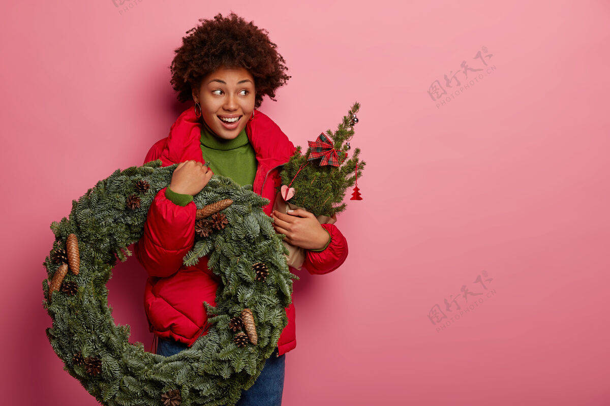 女人可爱的深色皮肤模特的摄影棚镜头带着圣诞花圈和装饰过的杉树 有节日的气氛 穿着红色外套 孤立在粉色背景上温暖姿势人